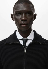 Czarny sweter na zamek z prążkowaną fakturą, z kołnierzem i białą koszulą pod spodem. Modelka ma neutralny wyraz twarzy.