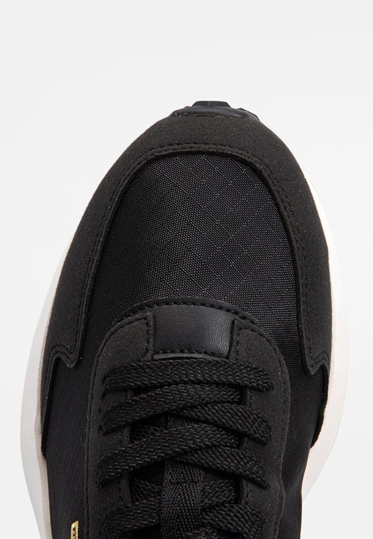 Zapatilla negra con parte superior de tela texturizada, que presenta una lengüeta acolchada, cordones planos negros y una suela de goma blanca. Detalles metálicos presentes.