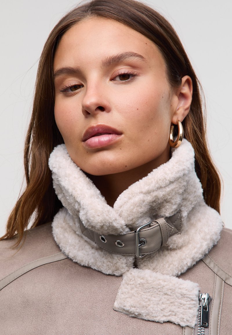 Collar de piel sintética en color crema con una correa gris que presenta herrajes metálicos. El material exterior tiene una textura suave en tono topo con detalles de cremallera.