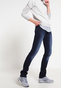 Navyblauwe slim-fit jeans, stretchdenim, gecombineerd met een grijze sweatshirt en grijze sport schoenen, die een gelaagde look met gestreepte details laat zien.