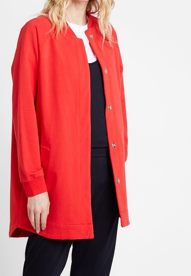 Femme portant une longue veste rouge boutonnée sur un haut blanc et un pantalon noir, debout sur un fond blanc uni.