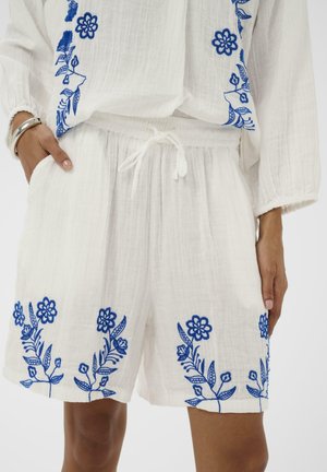 Witte loszittende korte broek met blauwe bloemige borduursels langs de zoom, gecombineerd met bijpassend topje, handen ontspannen langs het lichaam en in de zak.