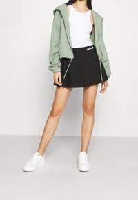 Světle zelená mikina na zip, bílý top bez rukávů, černá skládací sukně a bílé tenisky. Sukně má detail loga na pásku.