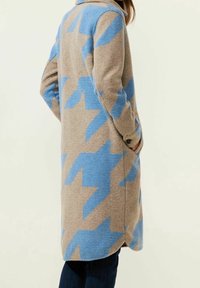 Lång kappa i beige tyg med djärva blå zickzackmönster, strukturerad krage och sidfickor. Mjuk textur och avslappnad passform.
