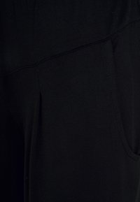 Schwarze Stoffhosen mit glatter Textur, die seitliche Taschen und flache Nähte für Komfort aufweisen. Der Stoff scheint dehnbar und figurbetont zu sein.