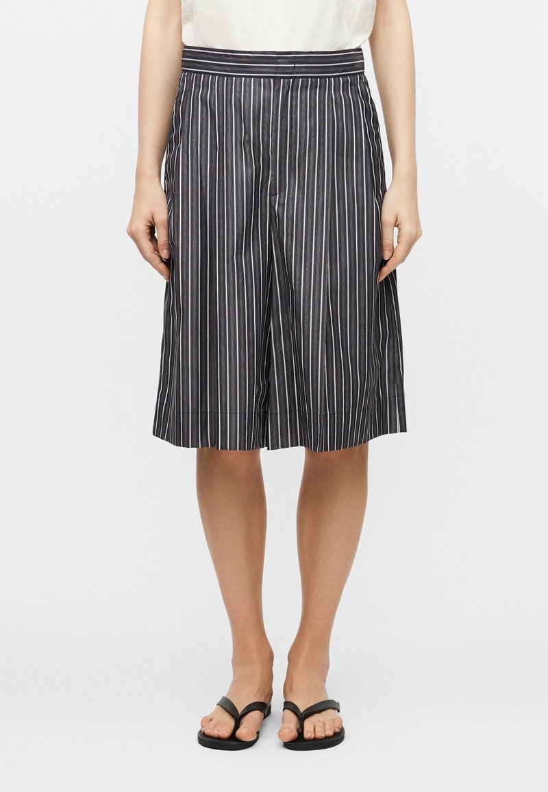 Γυναίκα που φοράει μαύρες και λευκές κάθετες ριγέ κολάν μέχρι το γόνατο με λευκό τοπ και μαύρα σανδάλια, στέκεται μπροστά σε απλό φόντο.