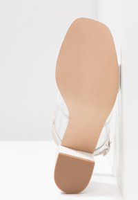 Sandale blanche avec une tige en plastique transparent et une semelle beige texturée et carrée. Le talon solide présente une légère brillance et une sangle de cheville ajustable.