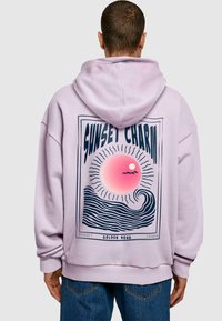 Personne portant un sweat à capuche violet clair avec un motif au dos représentant un soleil stylisé rose, des vagues noires et le texte "Charme du coucher de soleil Heure dorée".