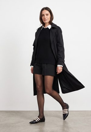 Kvinde iført sort trenchcoat, sort striktrøje, sorte shorts, gennemtrækkende strømpebukser og sorte sko med hvide stropper, stående på en grå gulv.