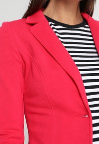 Blazer rose vif avec un col cranté et un détail de bouton unique, superposé à une chemise rayée noir et blanc. Texture de tissu lisse.