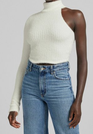 Valkoinen ribbineulottu pörröinen pooloneule, jossa on yksi pitkä hiha ja yksi hihaton puoli, yhdistetty korkeavyötäröisiin sinisiin farkkuihin.