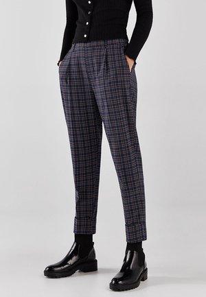 Personne portant un pantalon bleu marine à carreaux fuselé, des chaussettes courtes côtelées noires, des bottines en cuir noir brillant et un cardigan noir boutonné.