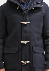 Uomo che indossa un cappotto toggle di un blu scuro sopra una camicia azzurra e un maglione di lana. Il cappotto presenta tre chiusure con corda e toggles in legno e due tasche frontali.