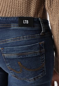 Vue arrière d'une personne portant un jean denim bleu avec une étiquette "LTB" et un pull en maille marron, montrant les détails de la poche et des passants de ceinture.