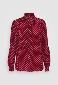 Blusa roja de manga larga con pequeño patrón de diamantes blancos, cuello alto y un gran lazo atado en el collar.