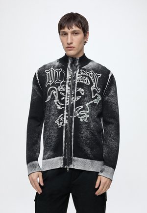 Jeune homme portant un pull zippé noir et blanc avec un grand dessin stylisé de dragon et un texte sur le devant, debout devant un fond uni.