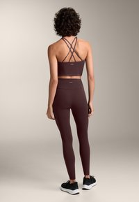 Bordo športni set, ki vključuje športni modrček z naramnicami in hlače z visokim pasom, narejen iz gladkega, raztegljivega materiala, v kombinaciji s črnimi športnimi čevlji.