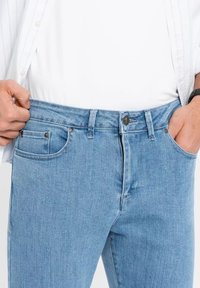 Muž v světle modrých džínách a bílé košili si stáhne pásek, aby ukázal volný střih v pase.