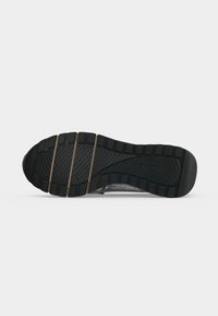 Semelle en caoutchouc noir d'une chaussure avec un motif de bande de roulement et trois rainures horizontales près des orteils, présentée sur un fond blanc uni.