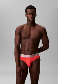 Rote Baumwollunterhose mit einem silbernen Bündchen, das das "Calvin Klein"-Logo trägt. Glatte Oberfläche und enge Passform, die die Körperform betont.