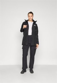 Black waterproof jacket with a zip front, adjustable hood, and logo. Paired with black cargo pants and black footwear. Smooth texture.