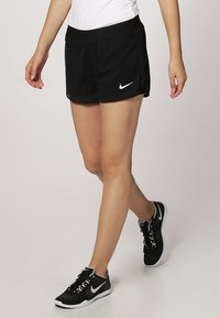 Persona che indossa pantaloni corti da corsa neri Nike e sneakers nere Nike, che cammina in avanti su uno sfondo uniforme.