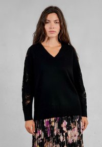 Maglione nero lavorato a maglia con scollo a V e lunghe maniche in pizzo; indossato con una gonna plissettata floreale caratterizzata da fantasie in viola, nero e beige.