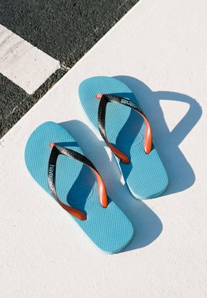 Par de chanclas azules con tiras negras y naranjas descansando sobre una superficie texturizada blanca y negra bajo la luz brillante del sol.