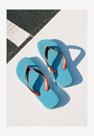 Ein Paar blaue Flip-Flops mit schwarzen und orangen Riemen liegen auf einer weiß-schwarzen strukturierten Oberfläche im grellen Sonnenlicht.