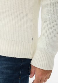 Maglione bianco lavorato a maglia con un motivo testurizzato, polsini e orlo a coste, con un piccolo etichetta del marchio sul lato, abbinato a jeans in denim scuro.