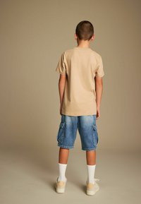 Camiseta de algodón beige, pantalones cortos cargo de mezclilla azul con bolsillos laterales, calcetas blancas hasta la rodilla y zapatillas de color crema. Vista trasera.