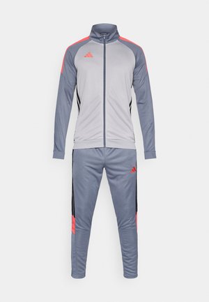 Sivi atletski trenirka z zadrgo, rdečimi poudarki in črnimi stranskimi črtami, združena z ujemajočimi se ozkimi hlačami.