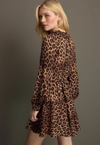 Leopardmönstrad klänning med långa ärmar, draperad halsringning och vågig kjol. Tillverkad av lättviktigt material i bruna och svarta toner.