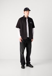 Zwarte short-sleeve button-up shirt over een wit long-sleeve shirt, gecombineerd met zwarte jeans en zwarte sneakers, afgemaakt met een zwarte pet.