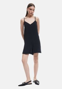 Sort ærmeløs jumpsuit med V-hals design, lavet af letvægtsstof. Kombineret med sorte flade sko med nitte-detaljer.