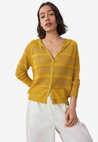 Gelber gestrickter Cardigan mit Kapuze, der horizontale Streifen, durchgehende Knopfschließung und gerippte Bündchen hat. Kombiniert mit weißen Hosen.