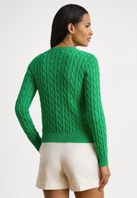 Grønn kabelstrikket genser med ribbet kant og mansjetter, med et teksturert design og tettsittende silhuett, kombinert med lyse shorts.