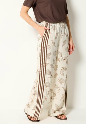 Persoon die losse crèmekleurige broek draagt met bruine schelpenprints en verticale bruine strepen, gecombineerd met een bruin topje en sandalen.