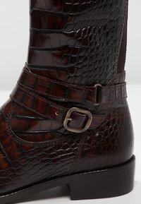 Bottes chelsea en cuir marron avec un motif crocodile, une bride décorative avec une boucle en métal, et un talon noir uni.