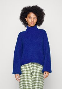 Maglione blu a collo alto in maglia con maniche ampie, abbinato a pantaloni a scacchi verdi e bianchi. Tessuto morbido, vestibilità rilassata e stile casual.