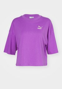 Fialové tričko s krátkým rukávem vyrobené z měkké tkaniny. Má kulatý výstřih a malé zlaté logo Puma na levé straně hrudi.