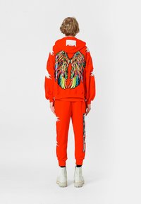 Sudadera roja con un diseño de alas multicolores y detalles desgastados, combinada con pantalones rojos a juego que presentan acentos gráficos y botas blancas.