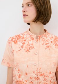 Lyhythihainen mekko, jossa on pyöreä kaulus, pehmeä persikanvärinen kangas, oranssin sävyinen kukkakuosi ja kolme etunappia.