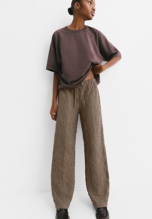Sudadera marrone oversize con scollatura rotonda abbinata a pantaloni taupe larghi e testurizzati, dotati di una cintura elastica.