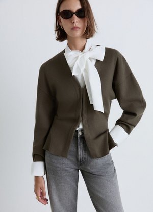 Donna che indossa occhiali da sole neri, camicetta bianca con grande fiocco, cardigan marrone e jeans grigi, in piedi con una mano sui fianchi.