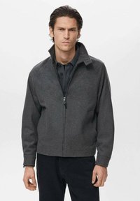 Šedá bunda s fleece podšívkou a vysokým golierom, s predným zipsom a postrannými vreckami; hladká štruktúra a priliehavý dizajn. Nosená cez tmavé tričko.