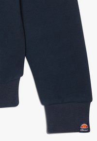 Manche de sweat-shirt bleu marine foncé avec poignet côtelé et petit patch logo de la marque Ellesse près du poignet.