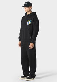 Sudadera negra con un diseño abstracto colorido en el lado izquierdo del pecho, combinada con pantalones de chándal negros a juego y zapatillas negras, todo en un ajuste casual.