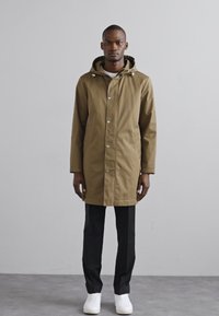Parka khaki resistente all'acqua con cappuccio, chiusura frontale a bottoni automatici e tasche laterali, indossata sopra pantaloni neri e sneakers bianche. Design semplice.