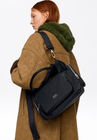 Ruda osoba w brązowym płaszczu z zielonym futrzanym kołnierzem, nosząca czarną torbę Bimba y Lola z srebrnymi zamkami na ramieniu.